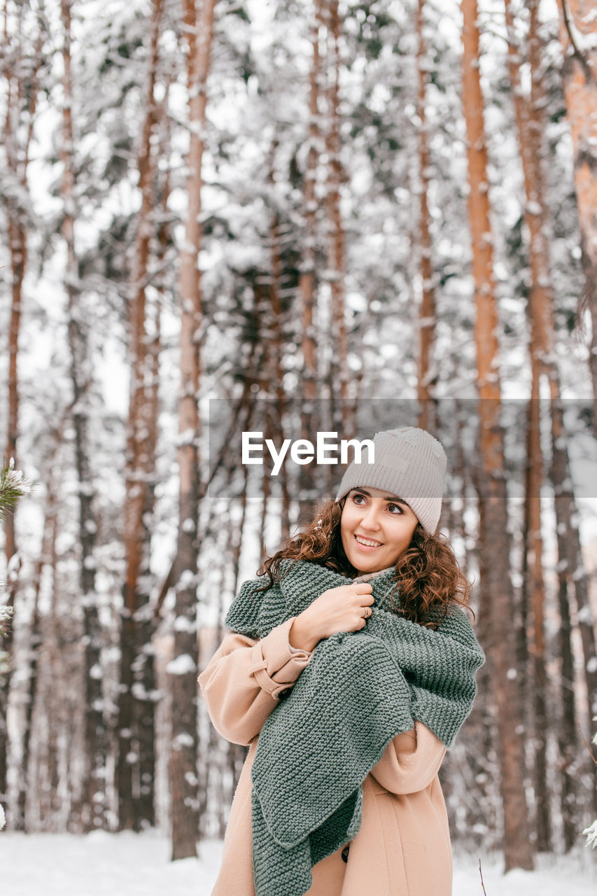 A smiling girl with a green scarf stands in a snowy forest in winter and looks away