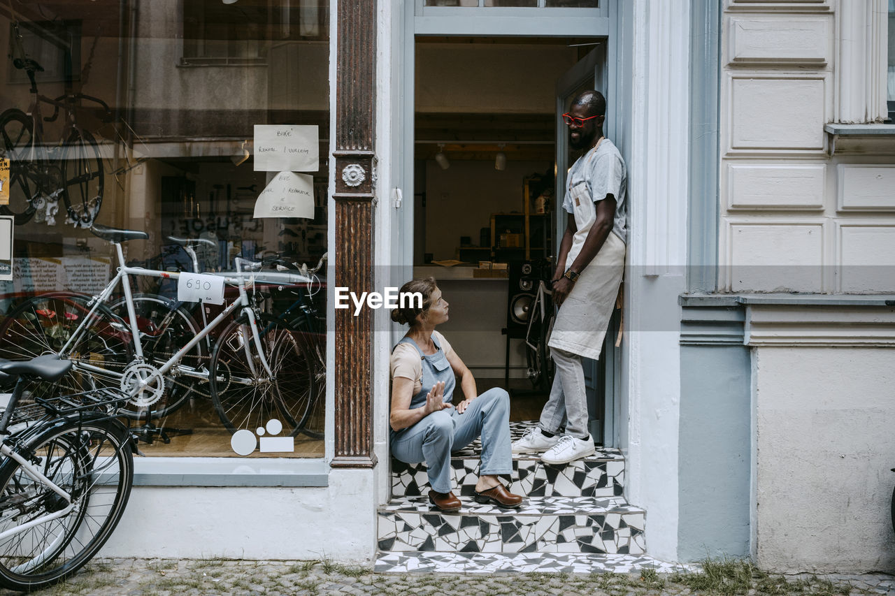 Senior owner talking to colleague while sitting on steps of bicycle shop