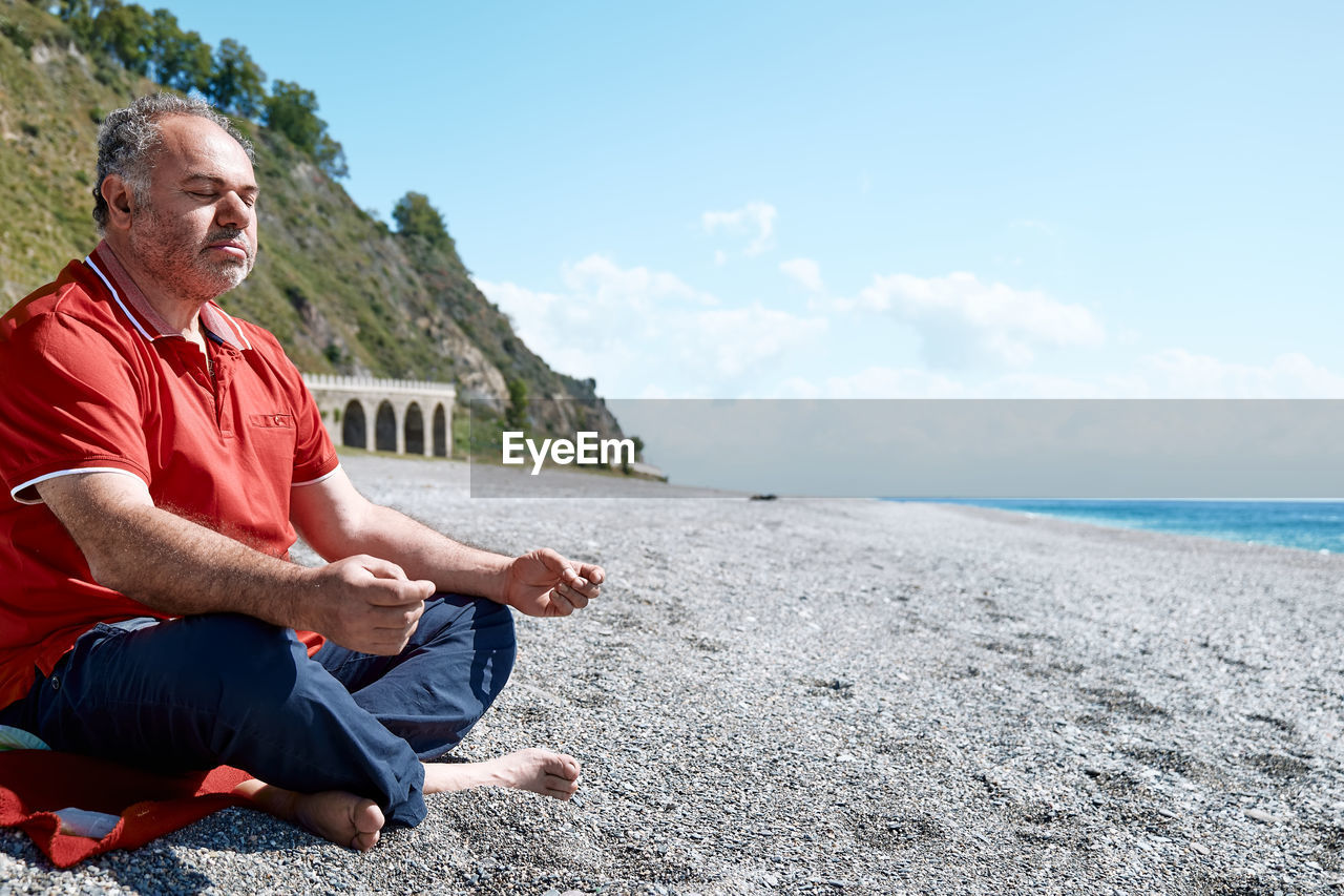 Full length of bearded man in casual clothes sitting in lotus pose on pebble beach and meditating