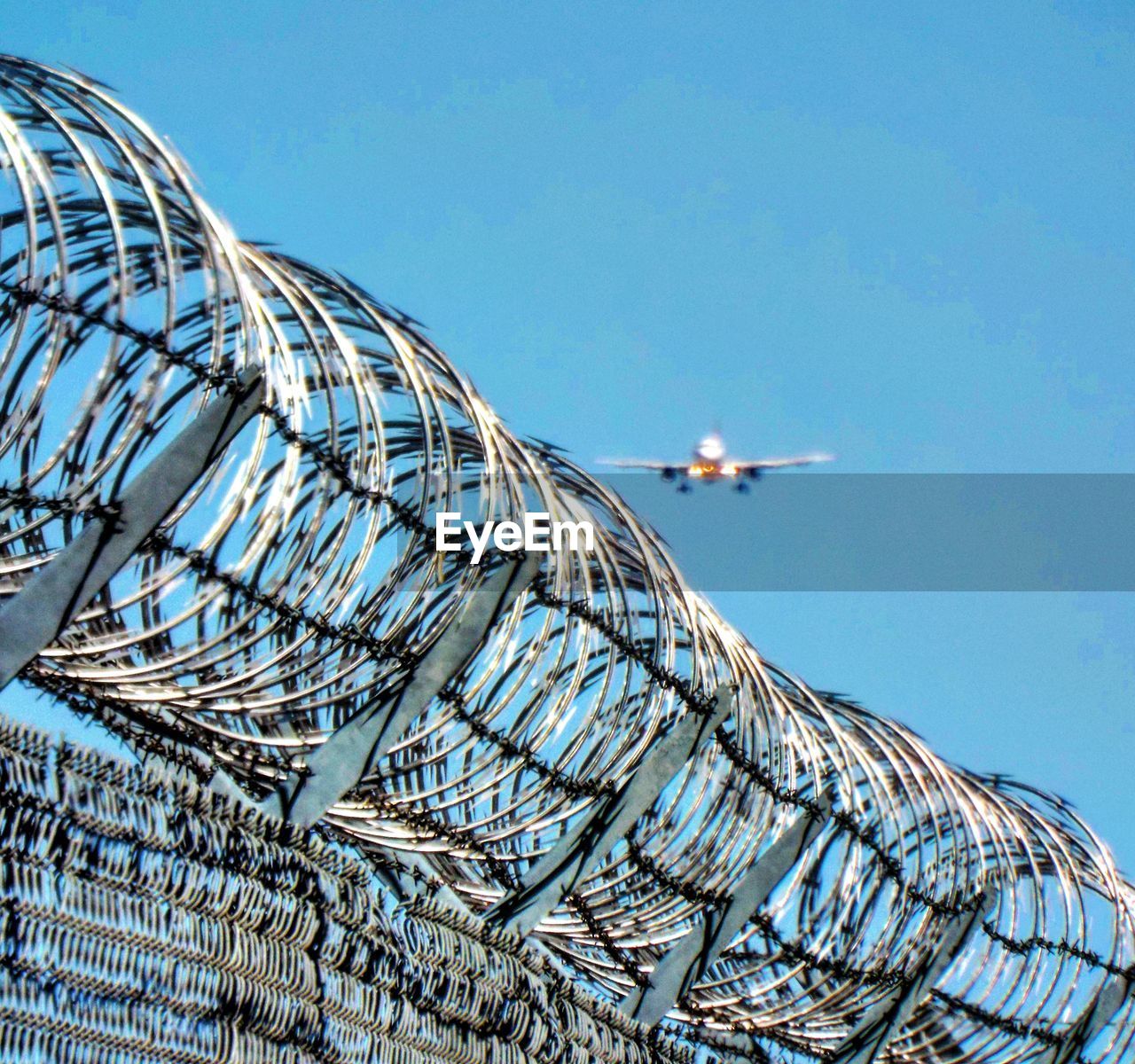 Low angle view of plane landing against clear blue sky with barbed wire fence 