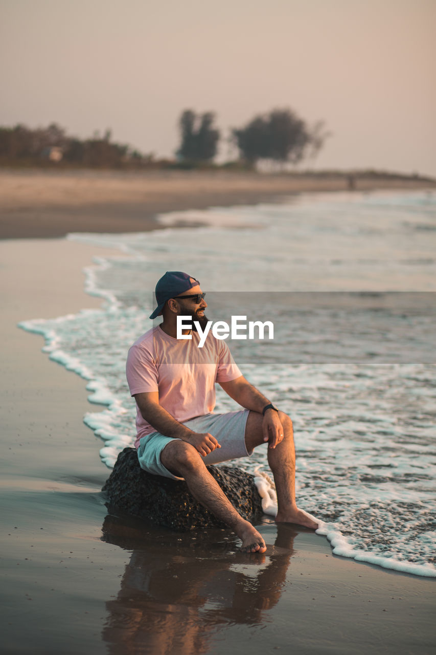 Side view of good looking bearded man sitting on beach during sunset with feet in the water