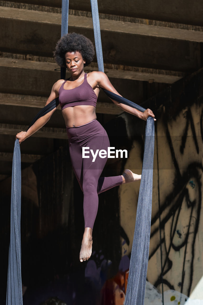 Slender african american female balancing on hanging hammock while doing aerial yoga above ground under bridge in city