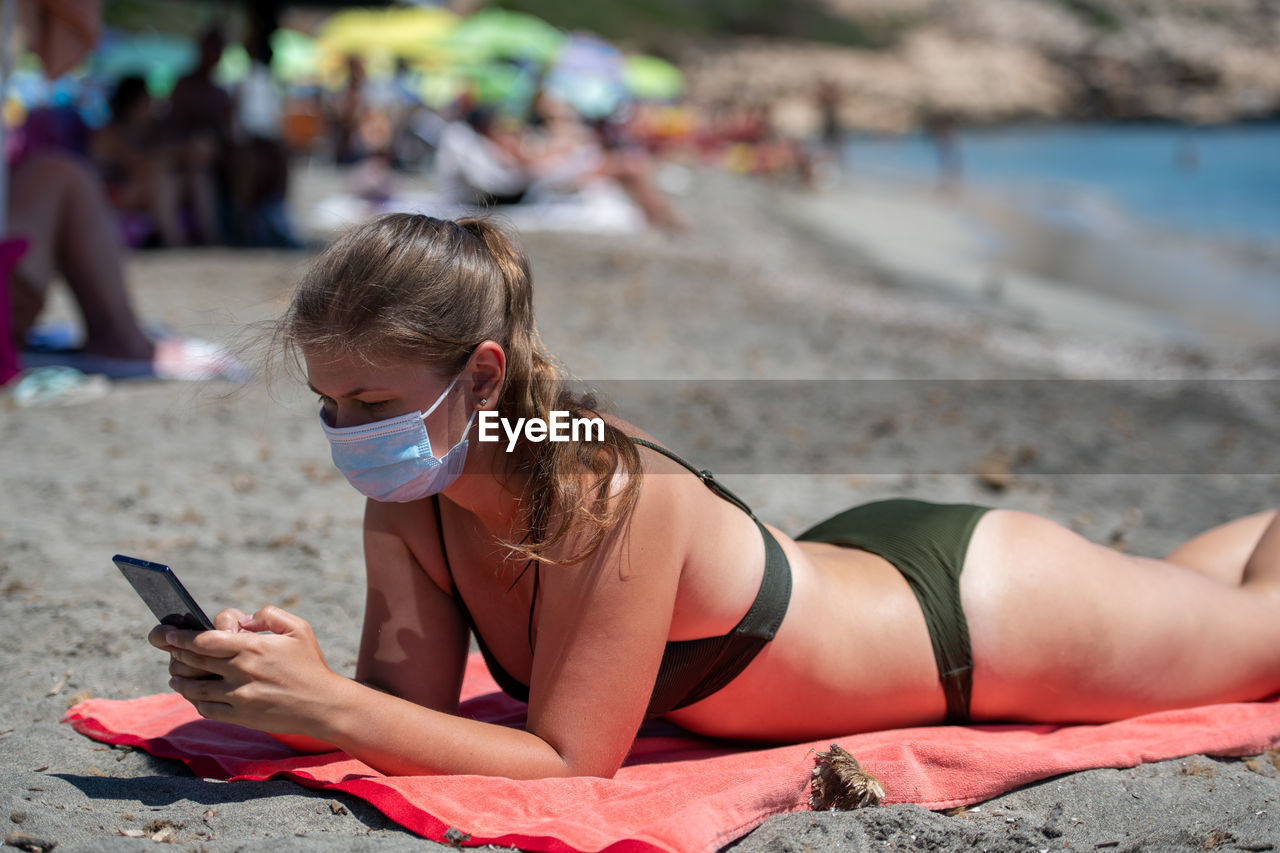 Woman wearing mask using mobile phone while lying on beach