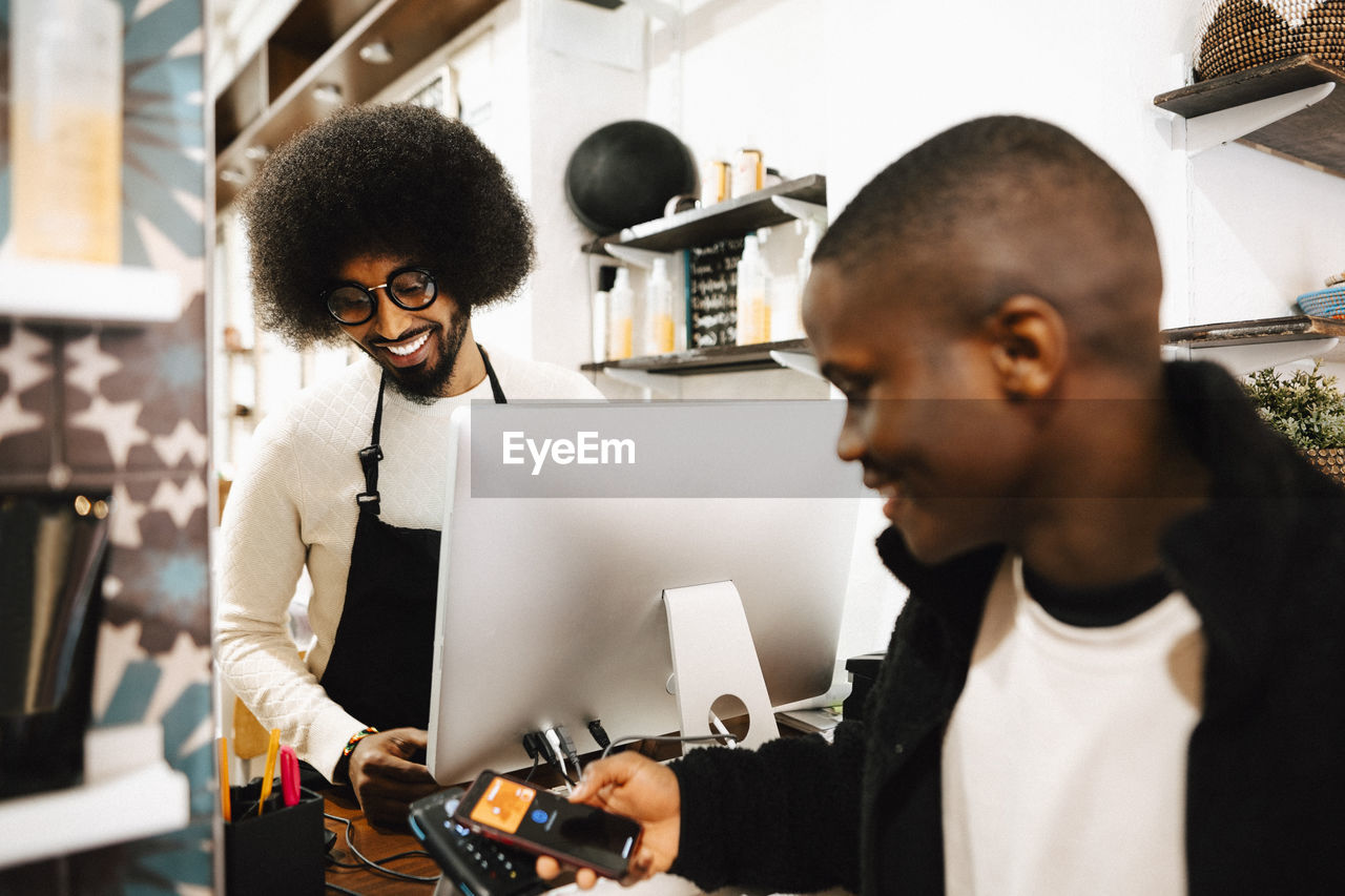 Smiling male customer doing contactless payment through smart phone at barber shop