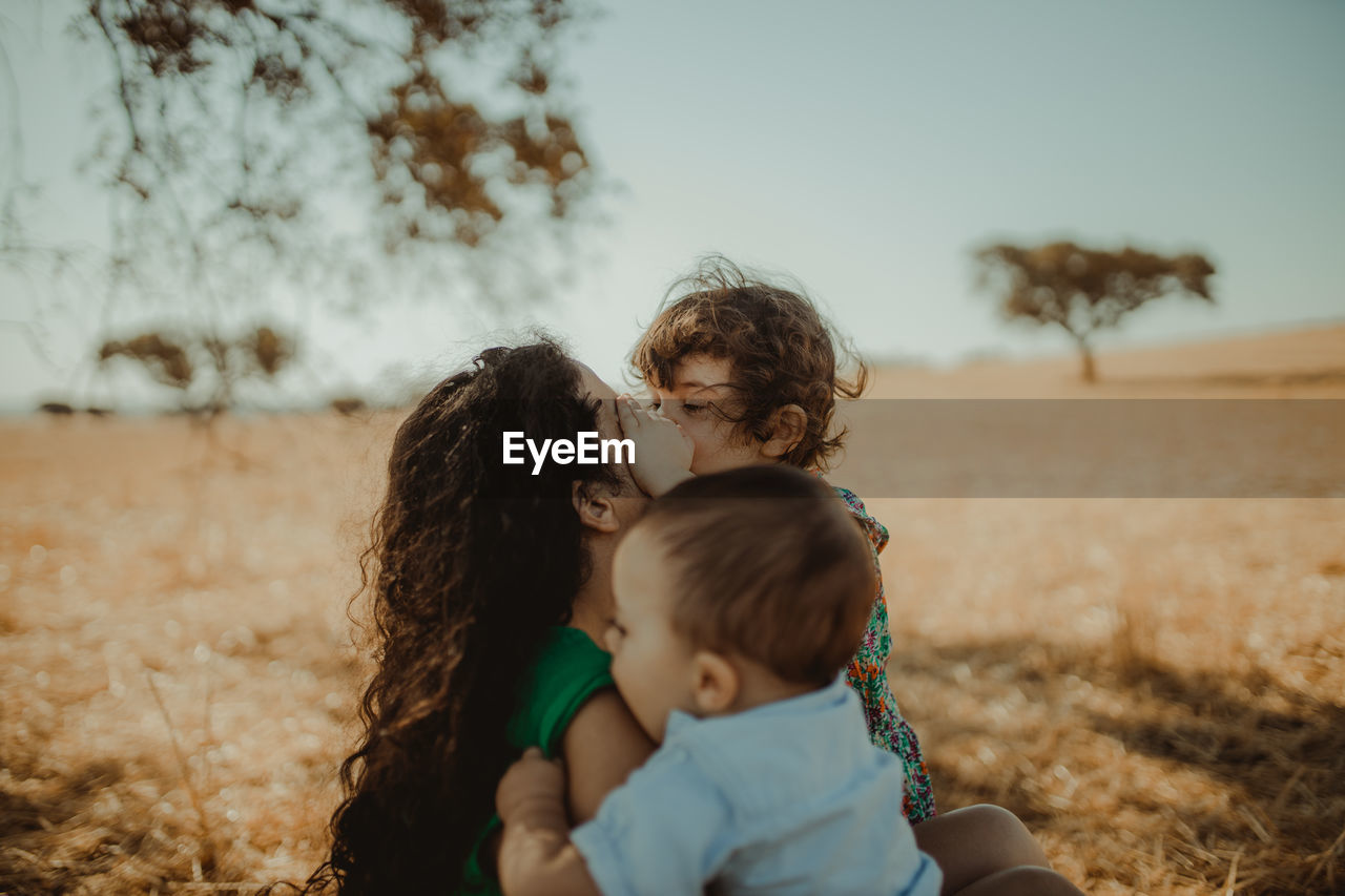 Mother and cute kids sitting on field against sky