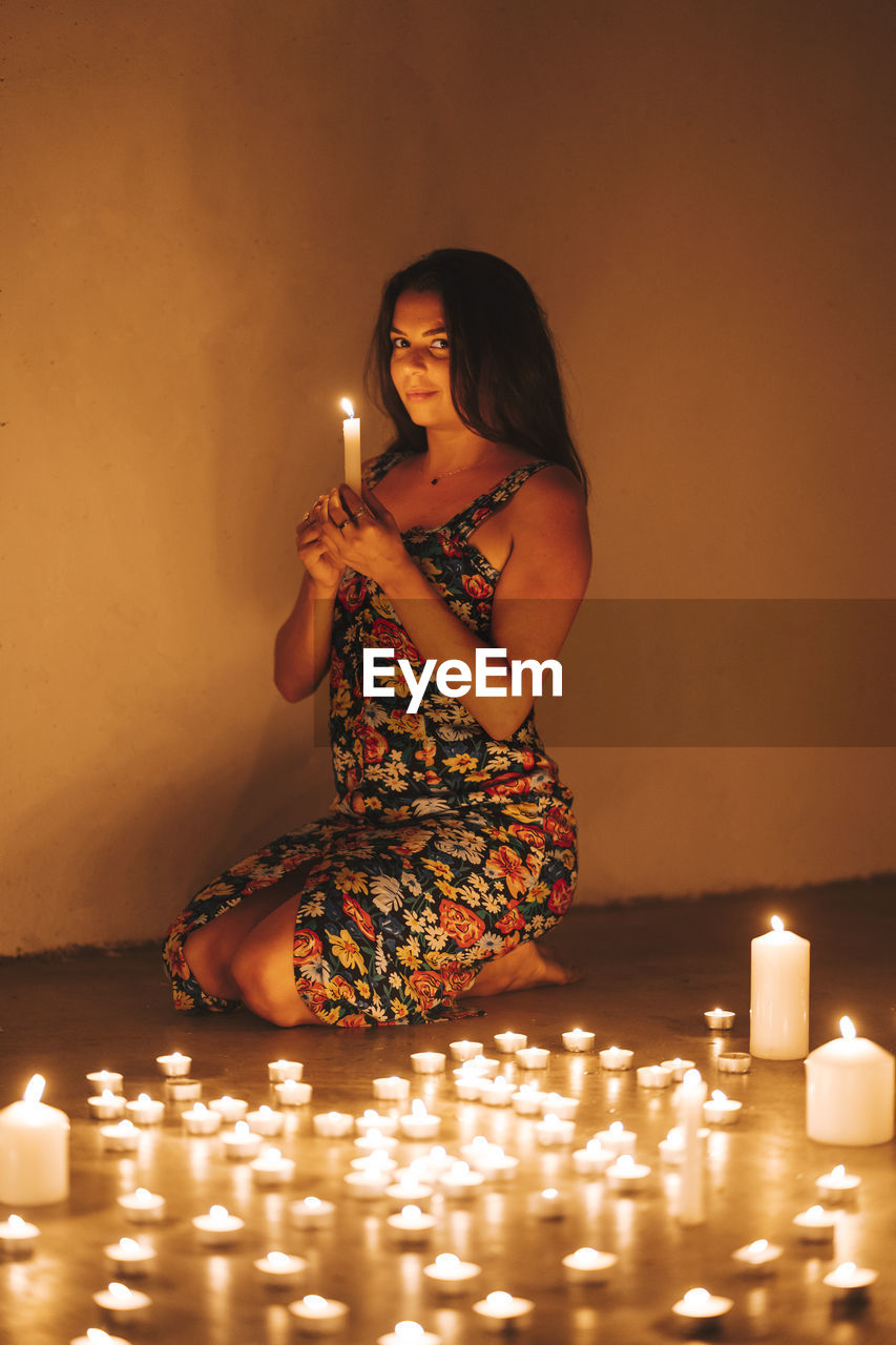 Woman with illuminated candles kneeling on floor by wall in darkroom