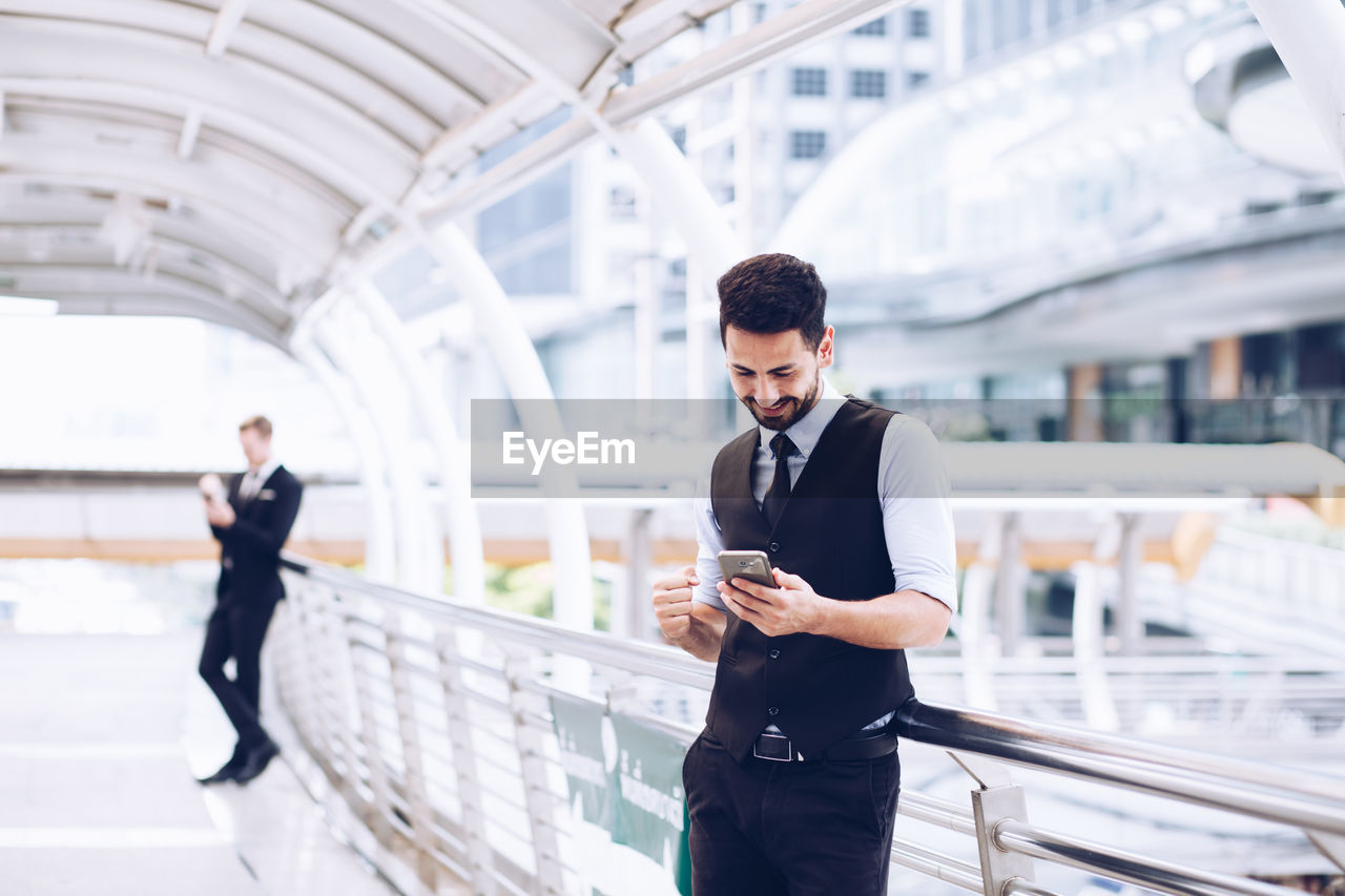 Smiling businessman using mobile phone while standing on bridge in city