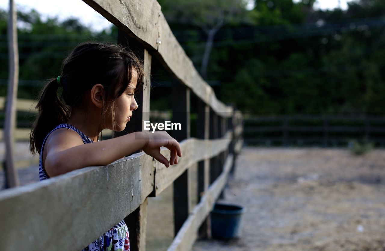 Side view of girl standing by fence at ranch