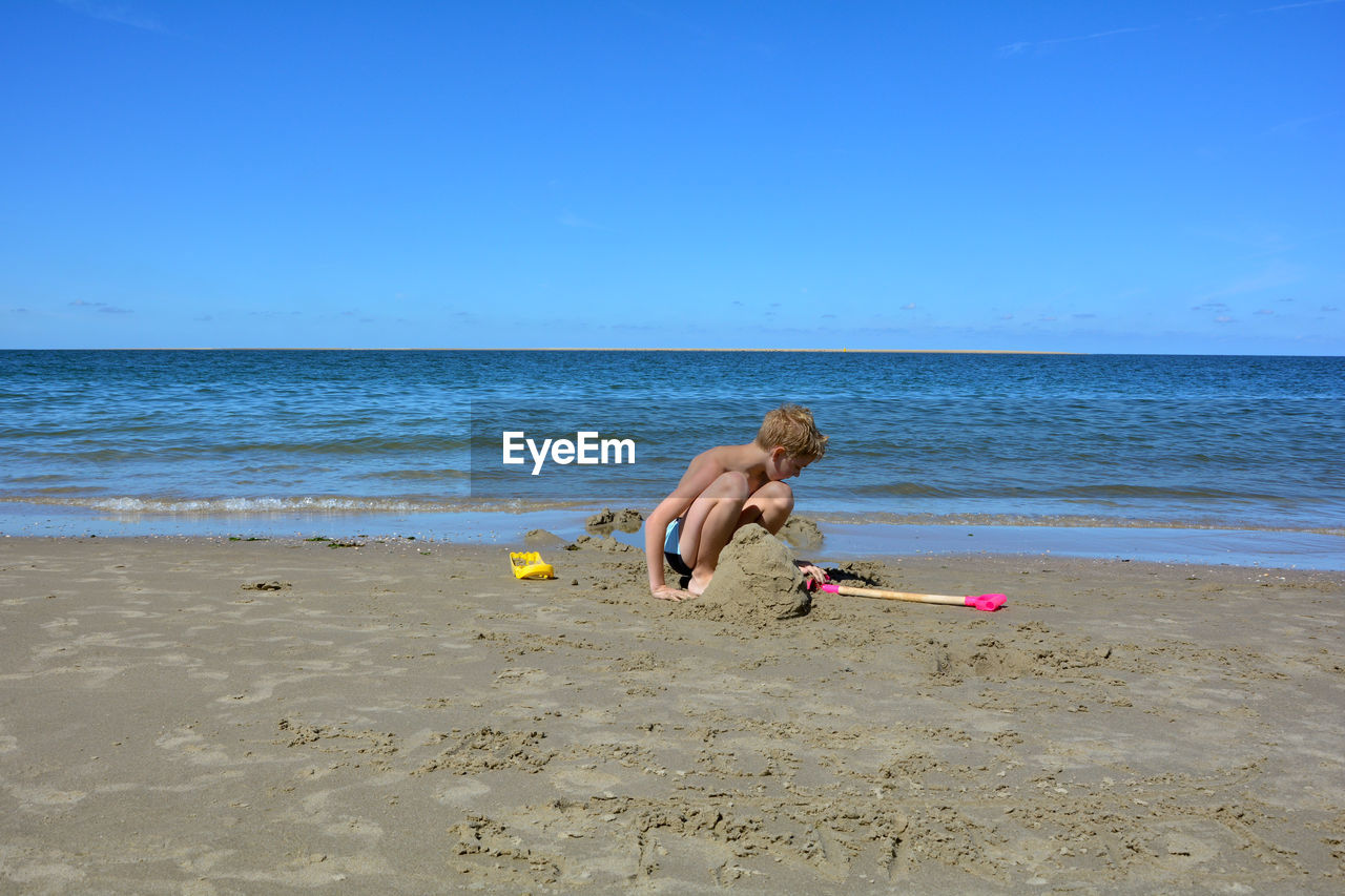 A blond boy with swimming trunks builds a sand castle in the sand on the beach in front of the sea