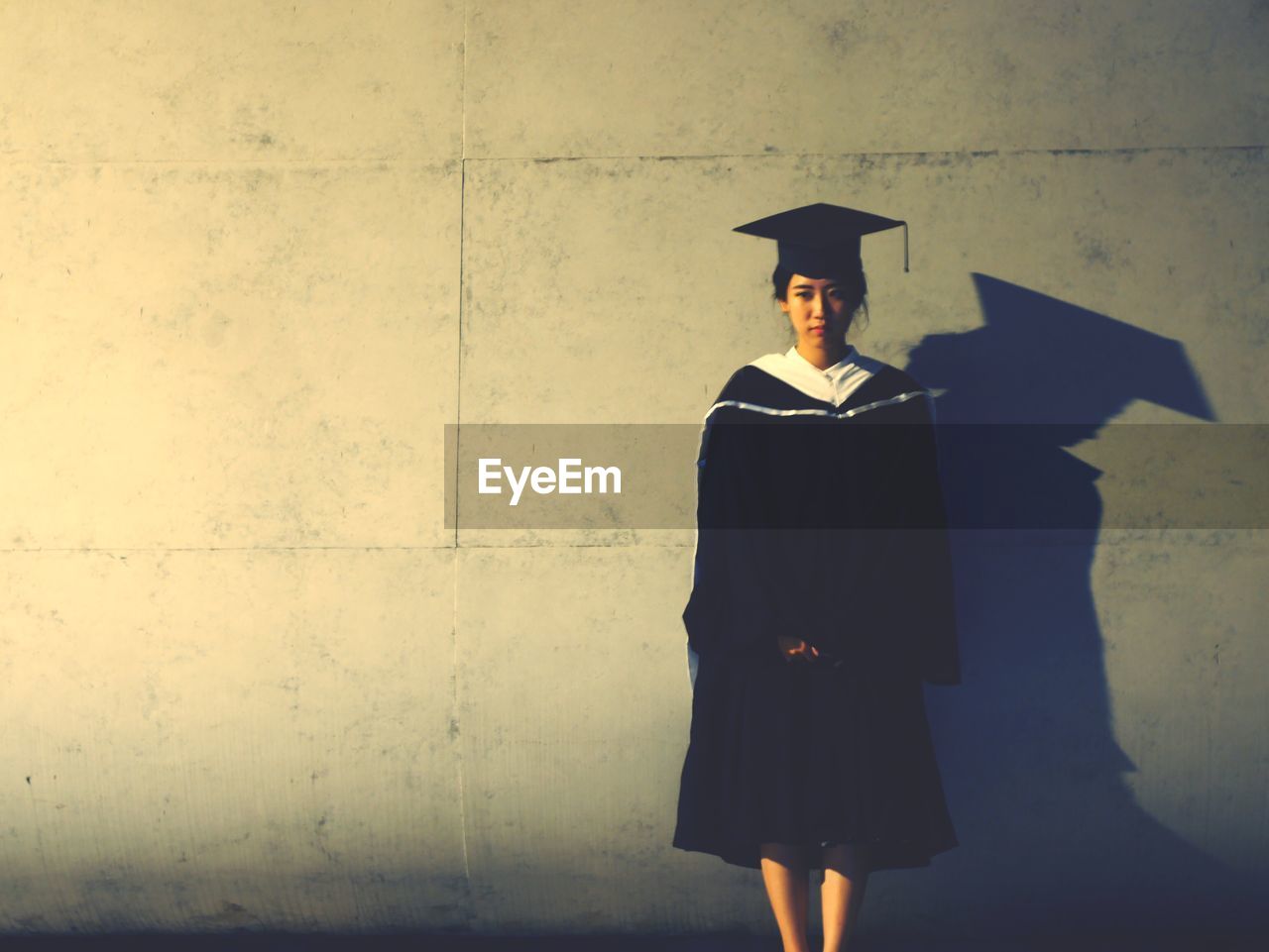 Portrait of woman in graduation gown standing against wall