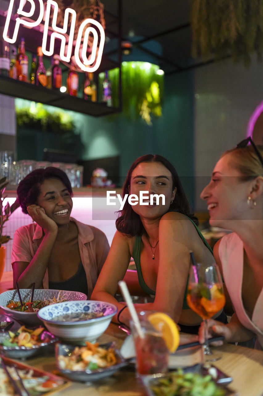 Smiling female friends talking with each other while sitting at illuminated restaurant