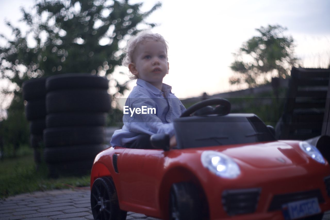 car, childhood, mode of transportation, motor vehicle, child, transportation, vehicle, one person, land vehicle, men, sports car, day, baby, portrait, nature, innocence, sitting, tree, looking, automobile, emotion, casual clothing, plant, front view, clothing, happiness, outdoors, looking away, toy, smiling, standing, toddler, supercar, cute, full length, driving, red