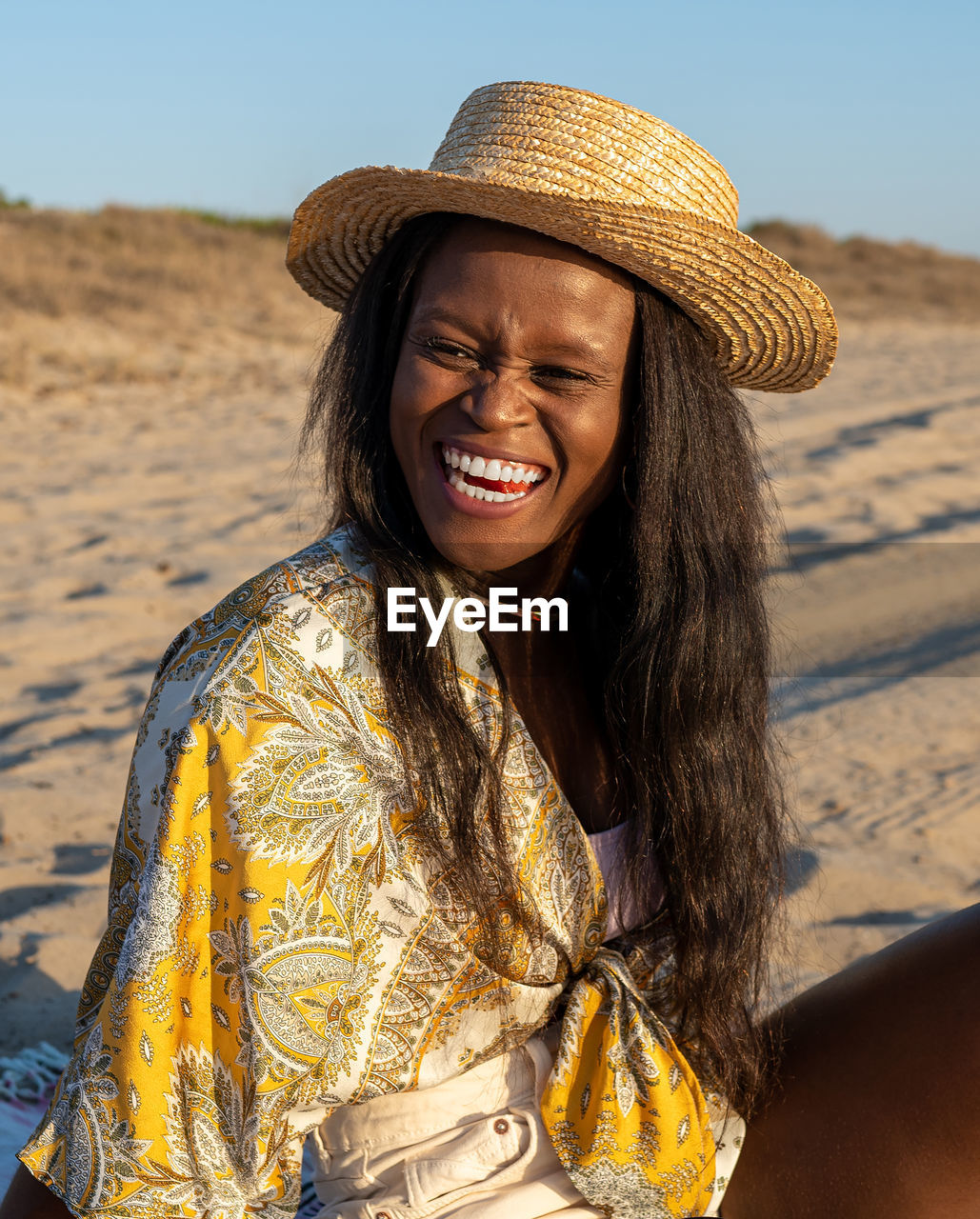 Cheerful african american female in beachwear and sunhat looking away and laughing at joke while spending sunny summer day on beach