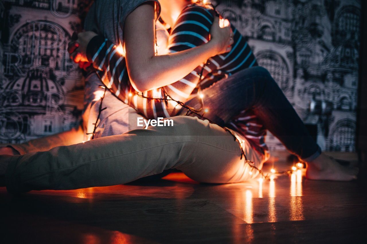 Low section of couple with illuminated string lights embracing at home