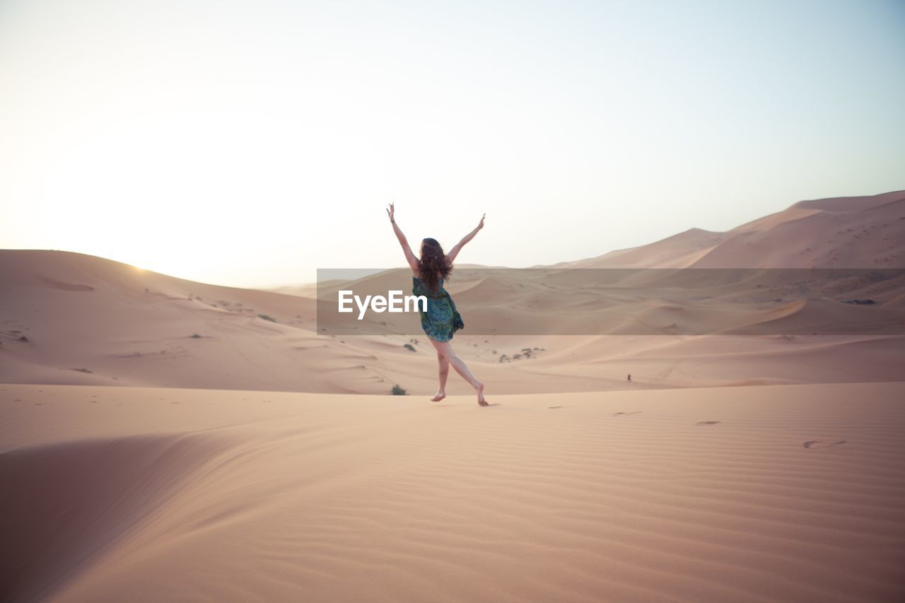 Rear view of excited woman with arms raised on sand dunes against clear sky