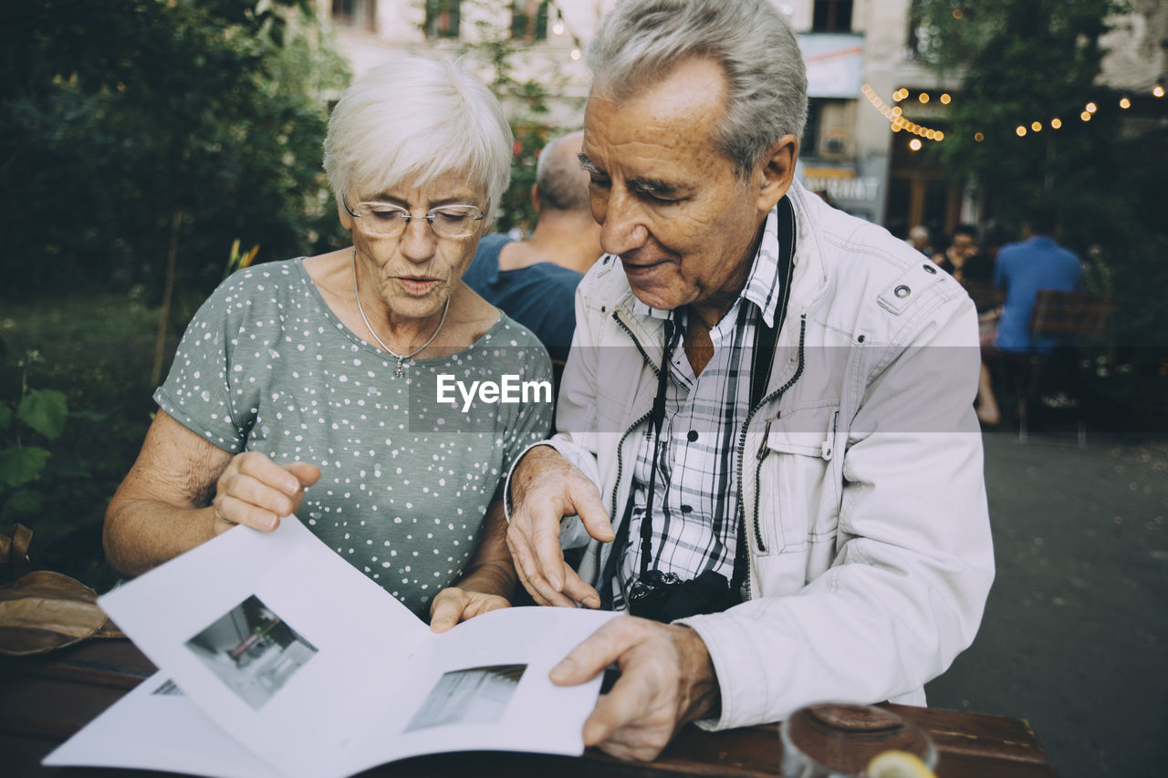 Senior man and woman tourist looking at pictures in book while sitting in restaurant