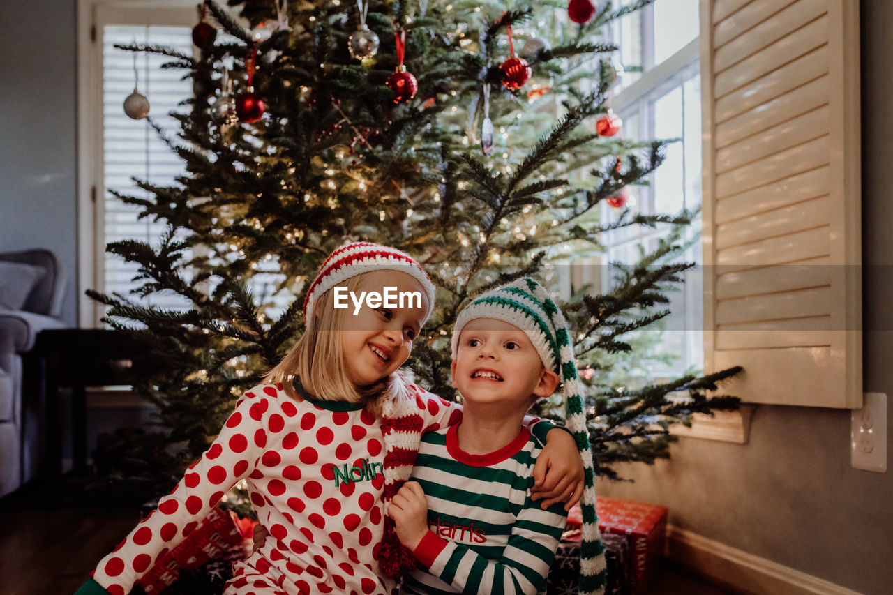 Siblings in costume sitting against christmas tree at home