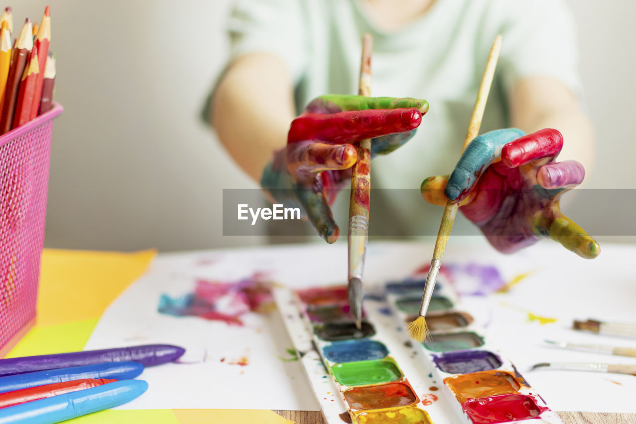 Cropped hand of boy holding paintbrush over palette while painting at classroom