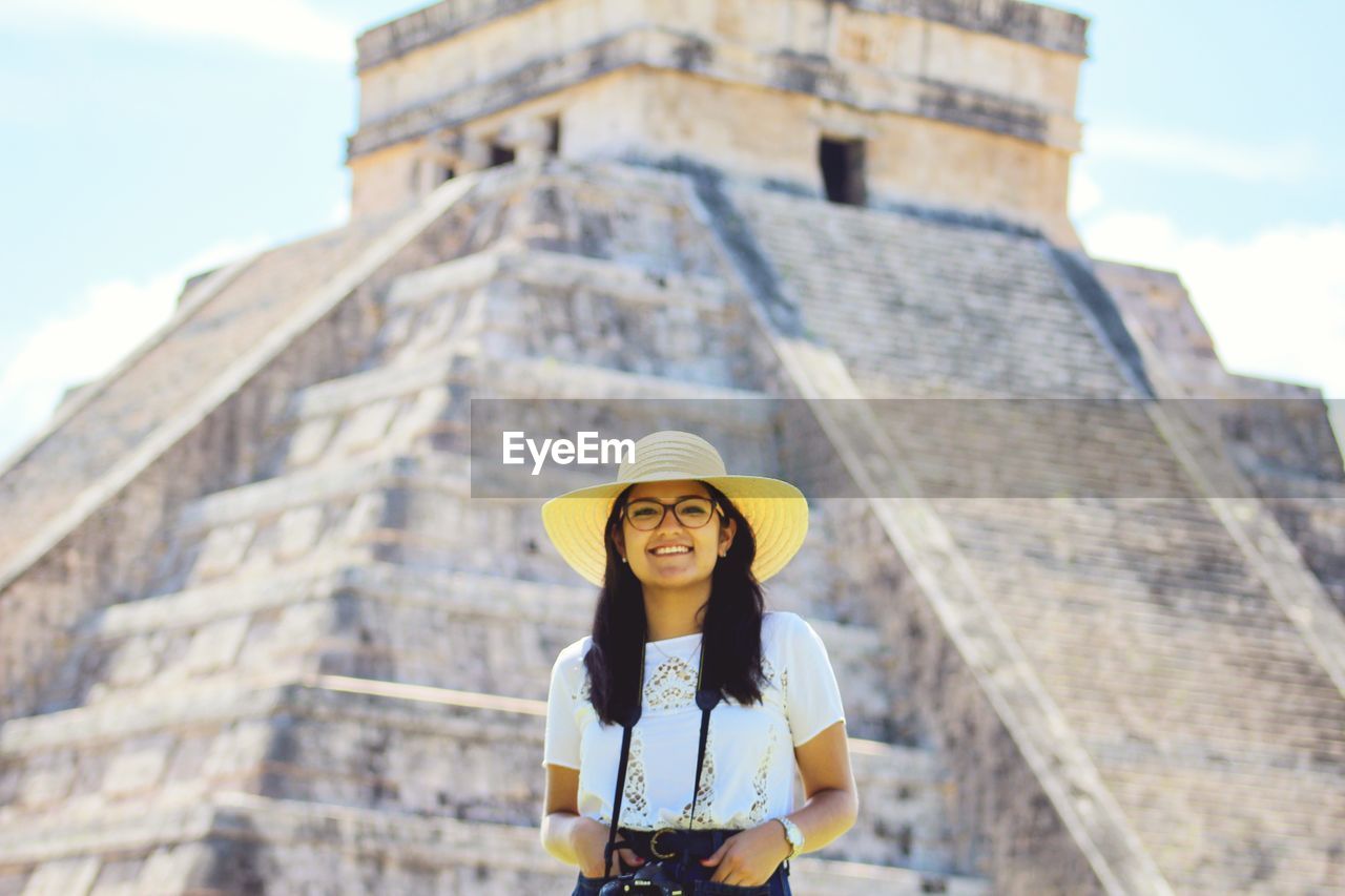 Portrait of smiling young woman wearing hat while standing against monument