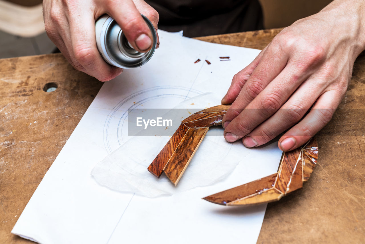 From above of crop anonymous male artisan applying adhesive spray on wooden piece on paper sheet in workroom