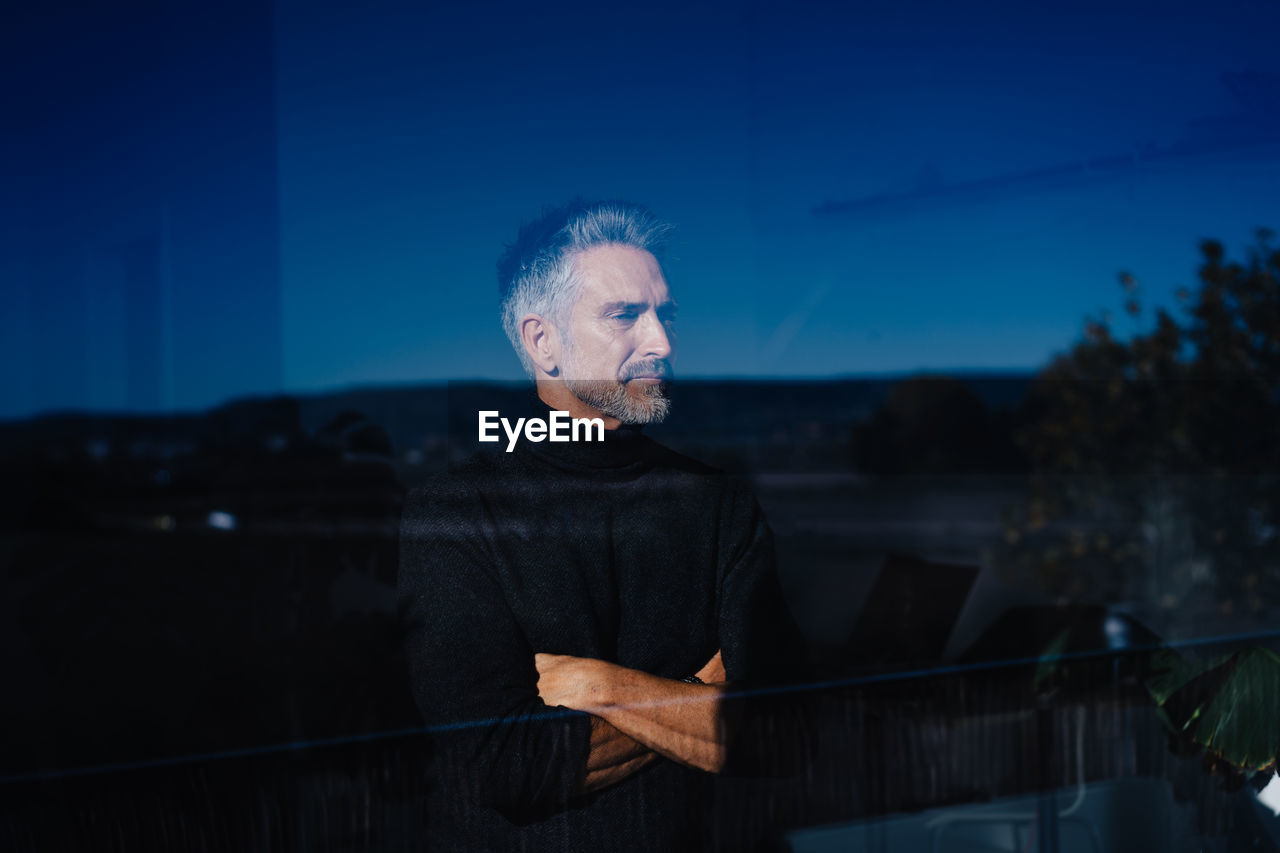 Gray haired senior male standing with crossed arms behind window reflecting landscape under cloudless sky in evening