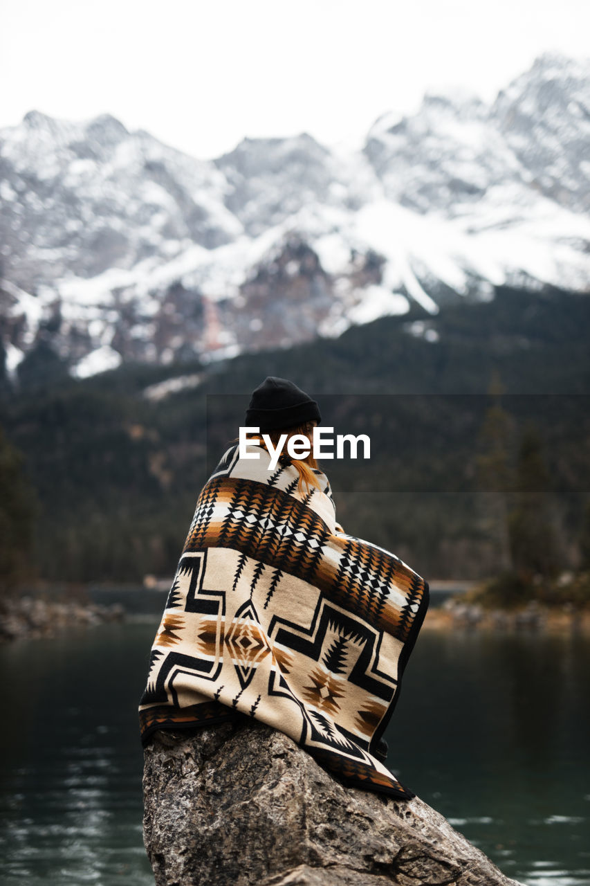 Side view of traveling female wrapped in blanket sitting on rock near pond in mountains and relaxing during vacation in winter