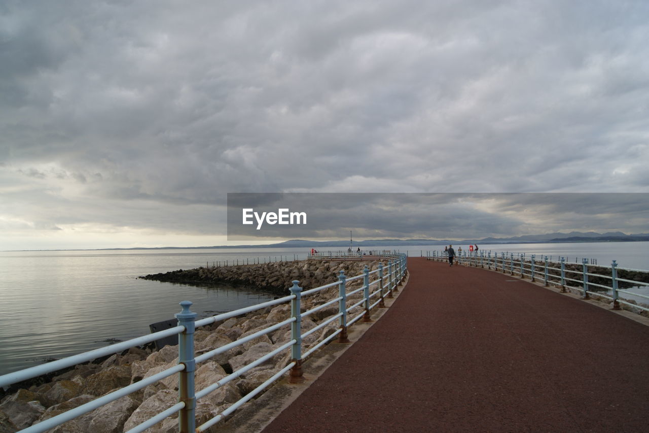 NARROW PATHWAY ALONG CALM SEA AGAINST CLOUDY SKY