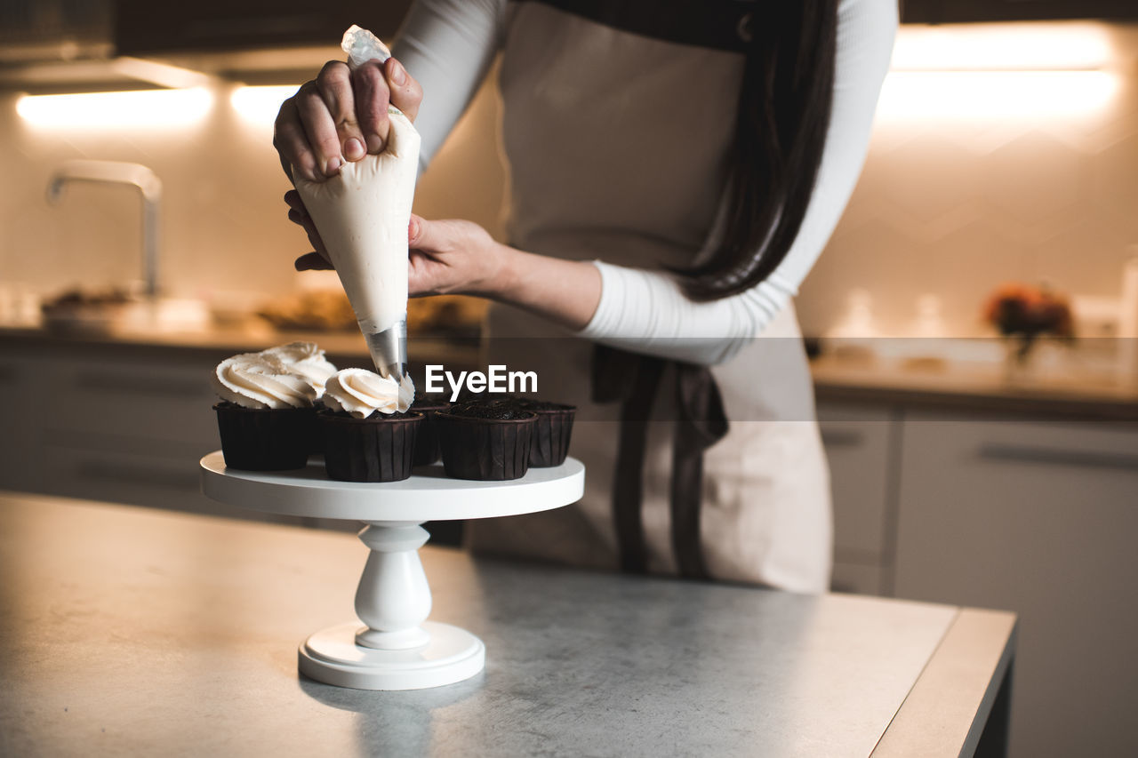 Woman decorate chocolate muffins with whipped cream cheese in kitchen table closeup. 