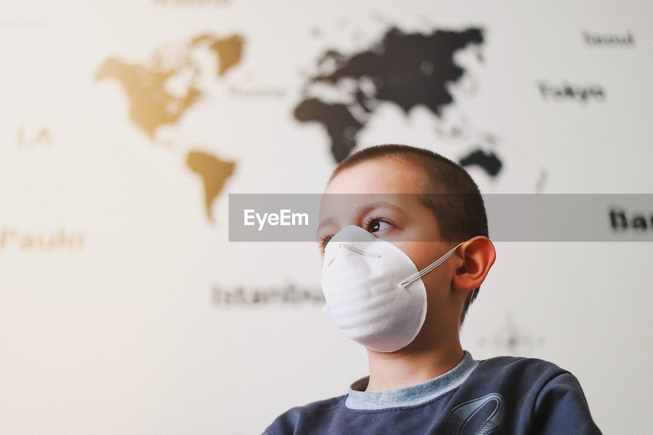 Close-up portrait of boy wearing a protective mask standing against wall with a world map detail