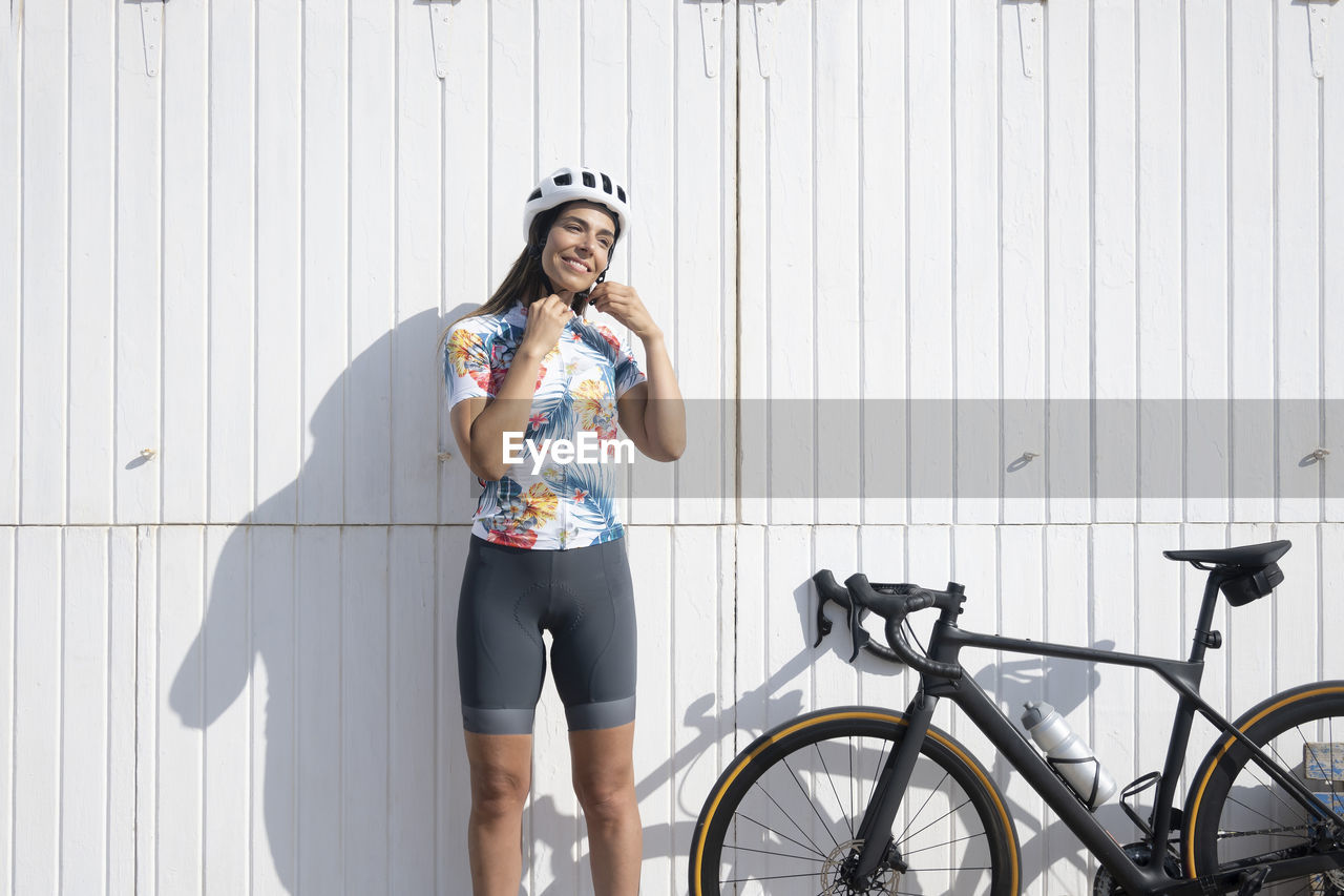 Happy cyclist adjusting helmet standing in front of white wall on sunny day