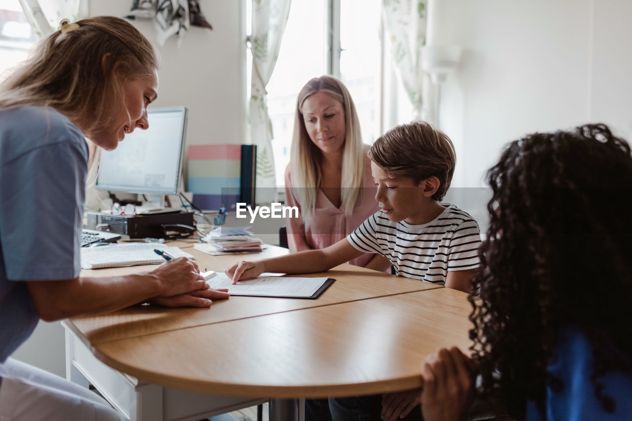 Doctor looking at patient pointing on paper while female nurse and mother sitting by desk in clinic