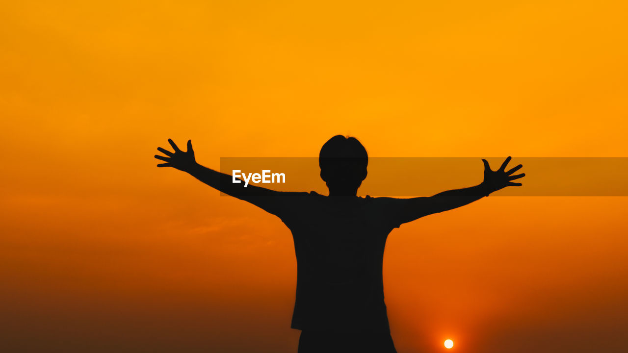 Silhouette man with arms outstretched standing against orange sky