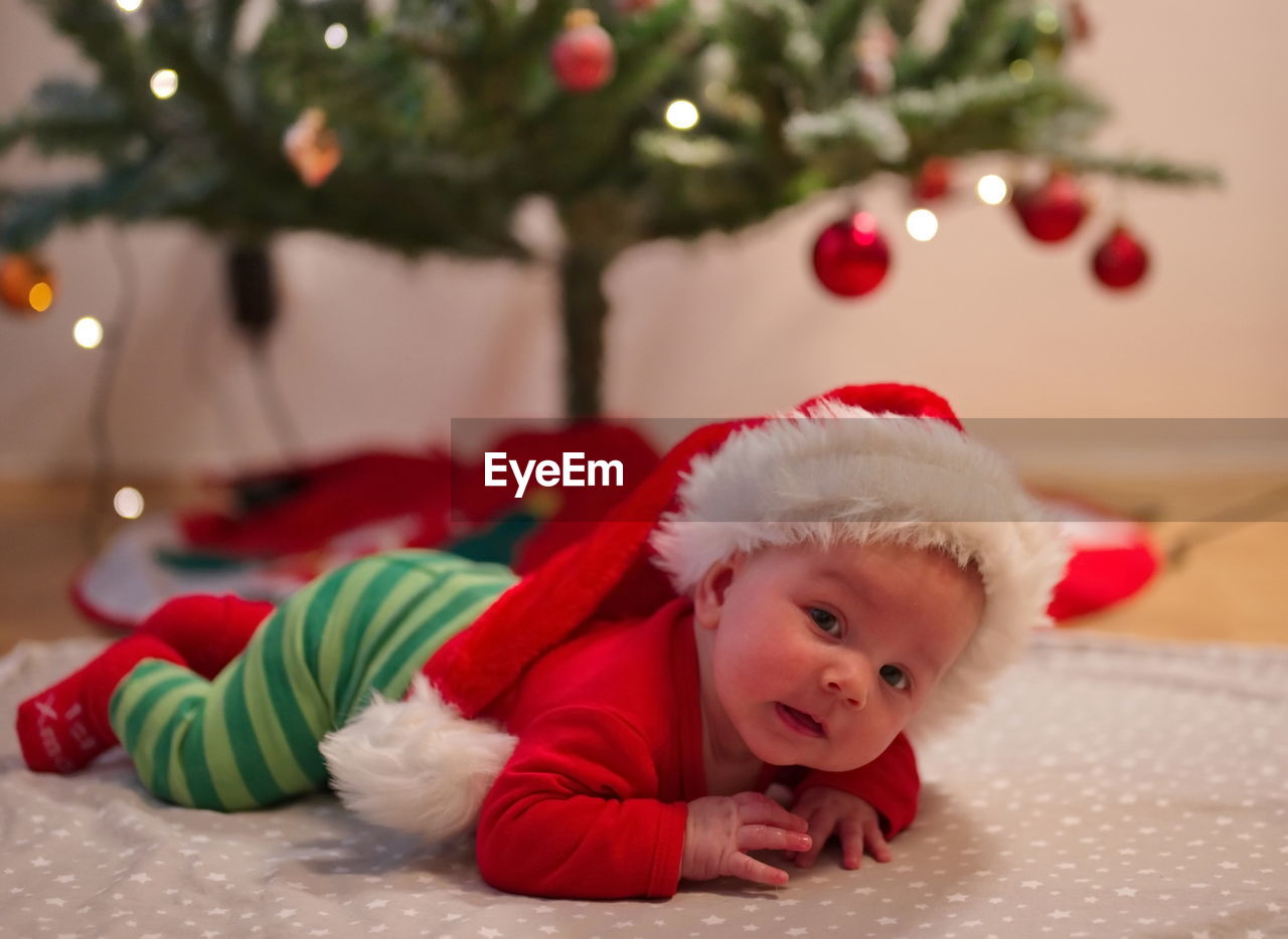 Newborn baby in santa clothes and christmas hat, lying on the ground next to christmas tree