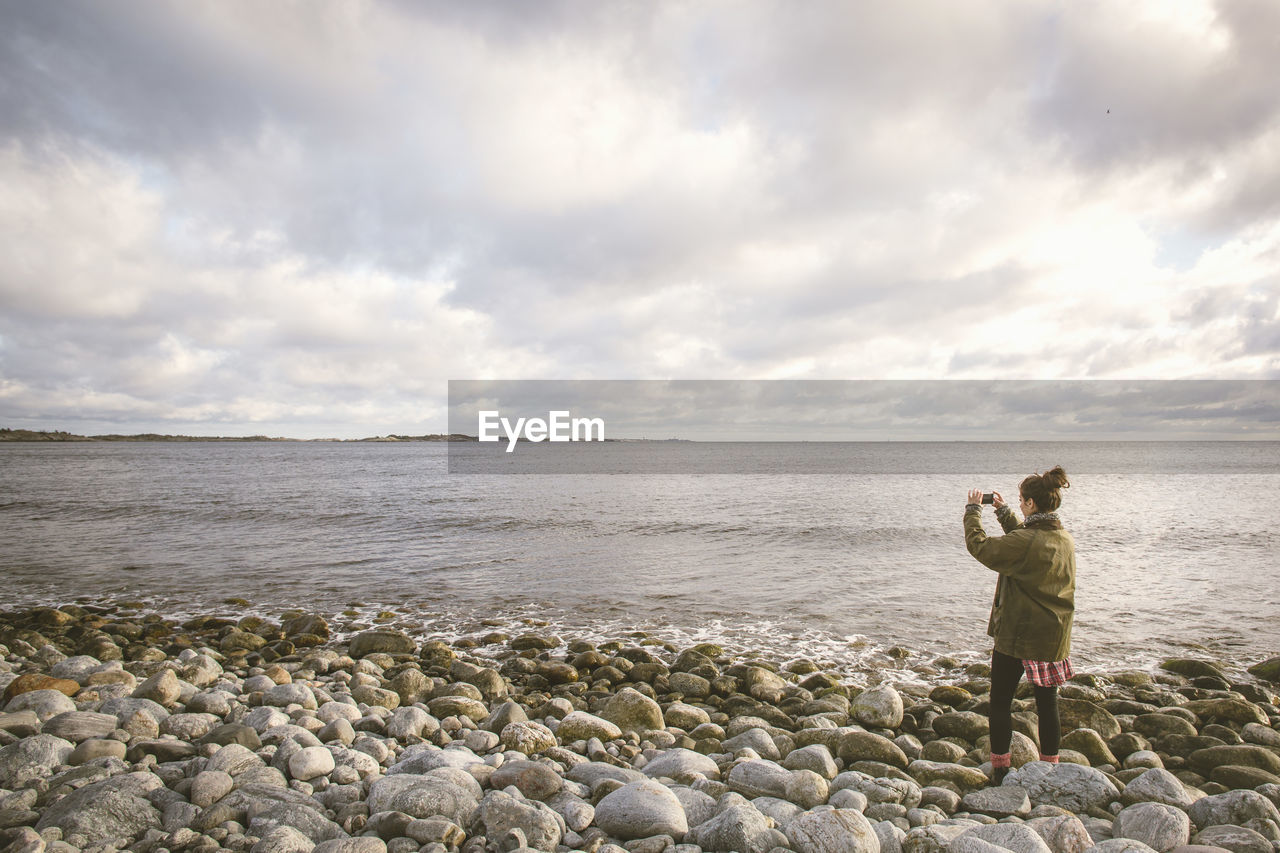 Woman photographing sea against cloudy sky while standing on shore at beach