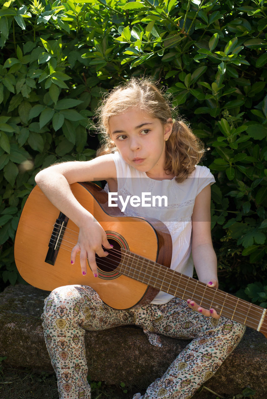 Girl playing guitar while sitting on rock against plants