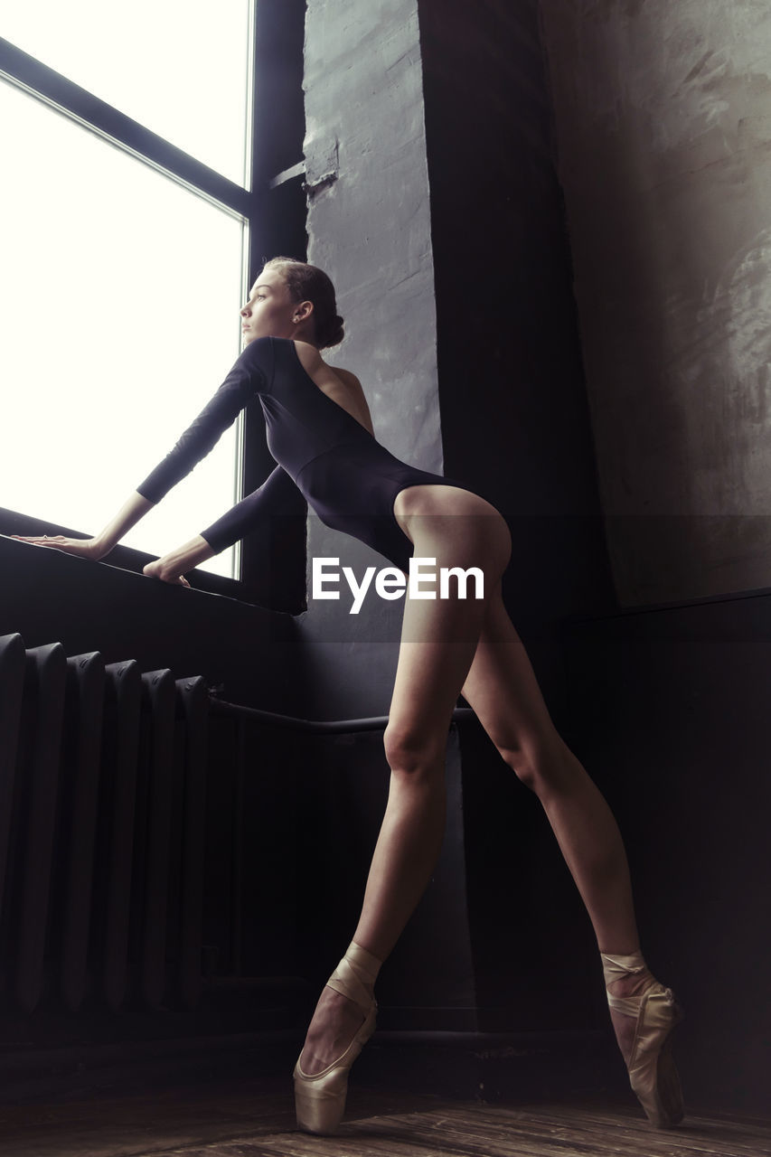 A ballerina in a bodysuit and pointes is standing in the corner of the room looking out the window