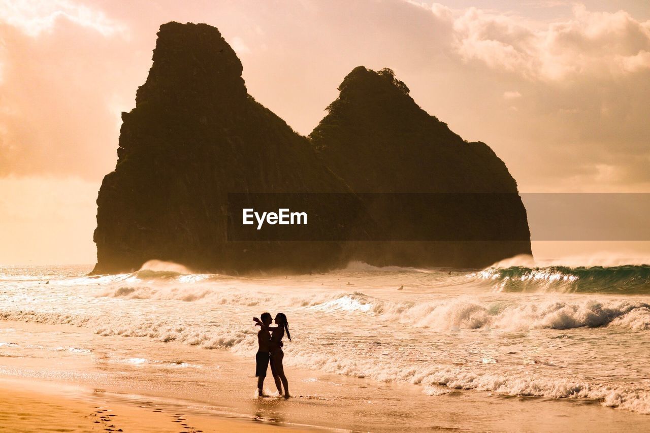 Couple embracing while standing at beach against rock formations during sunset
