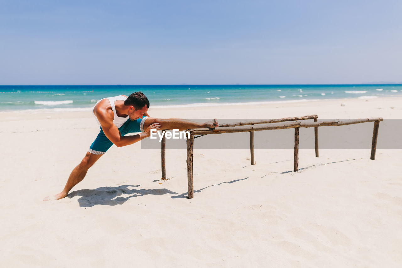 Man exercising on parallel bars at beach against blue sky