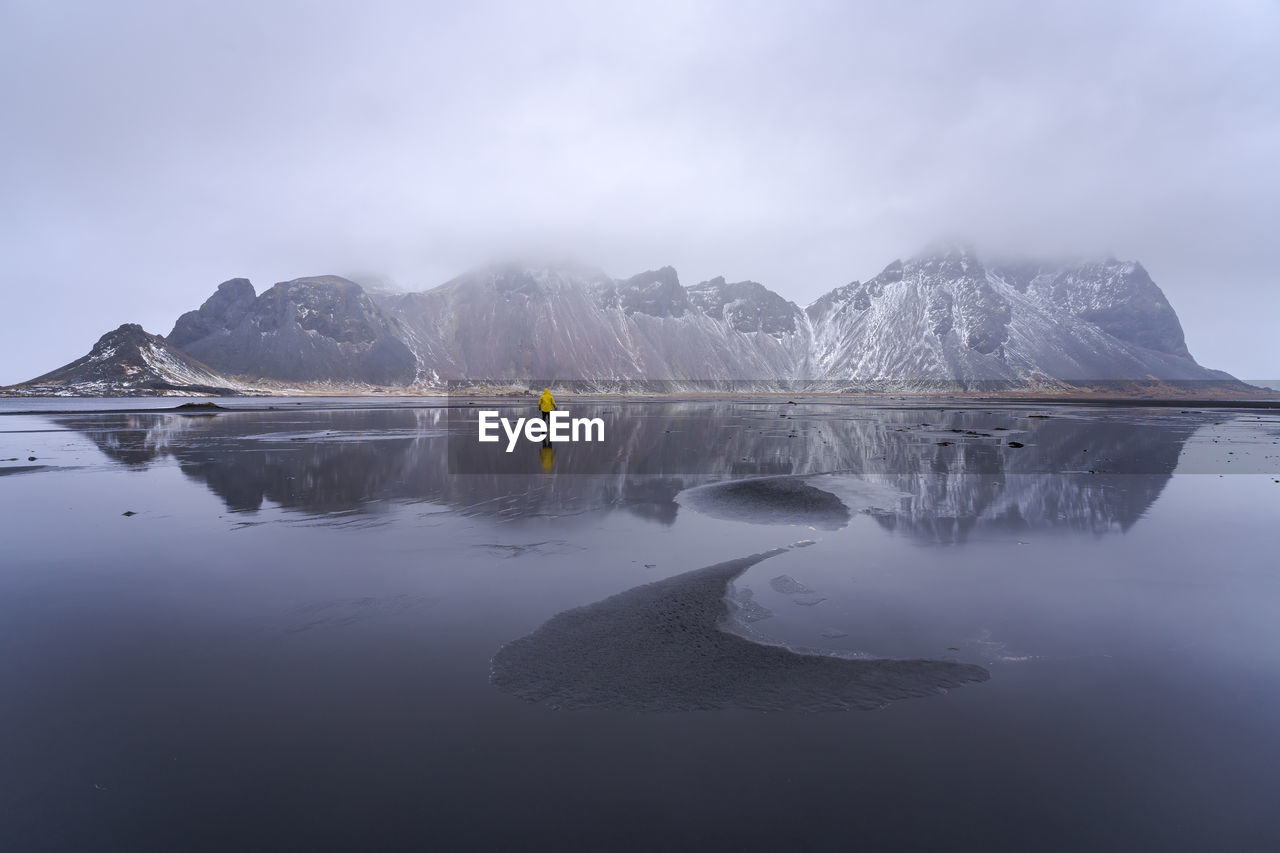 Distant view of unrecognizable traveler standing on wet black sand beach near waving sea against mountain ridge and cloudy sky in iceland