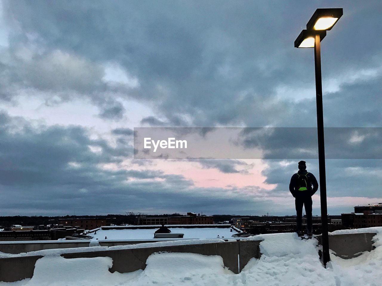 MAN STANDING ON SNOW COVERED FIELD AGAINST SKY DURING SUNSET