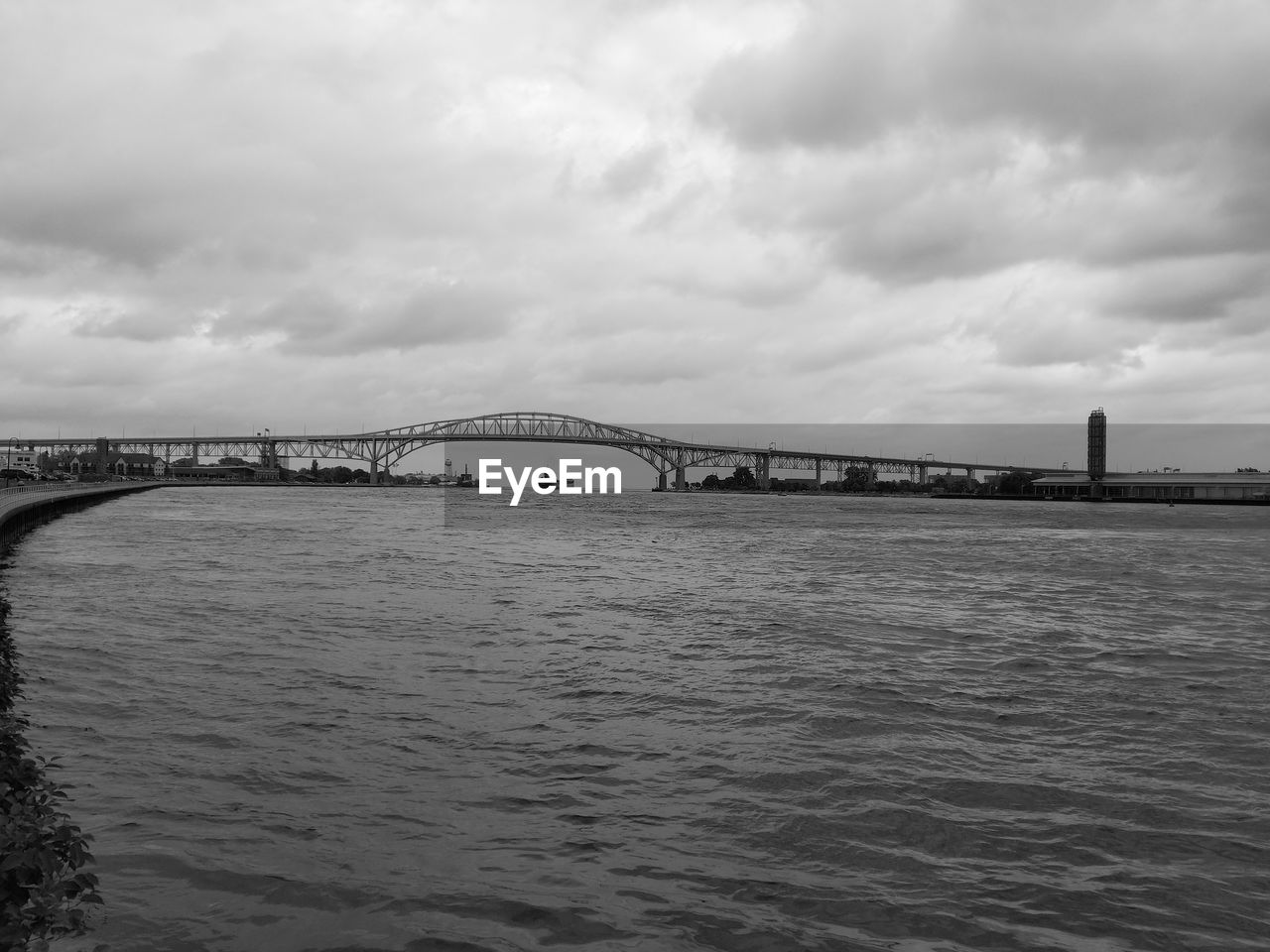 BRIDGE OVER CALM RIVER AGAINST CLOUDY SKY