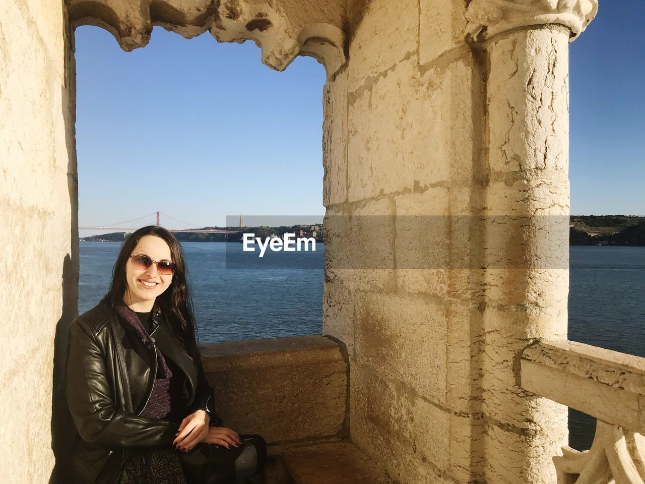 Portrait of smiling woman in sunglasses sitting against sea during sunny day