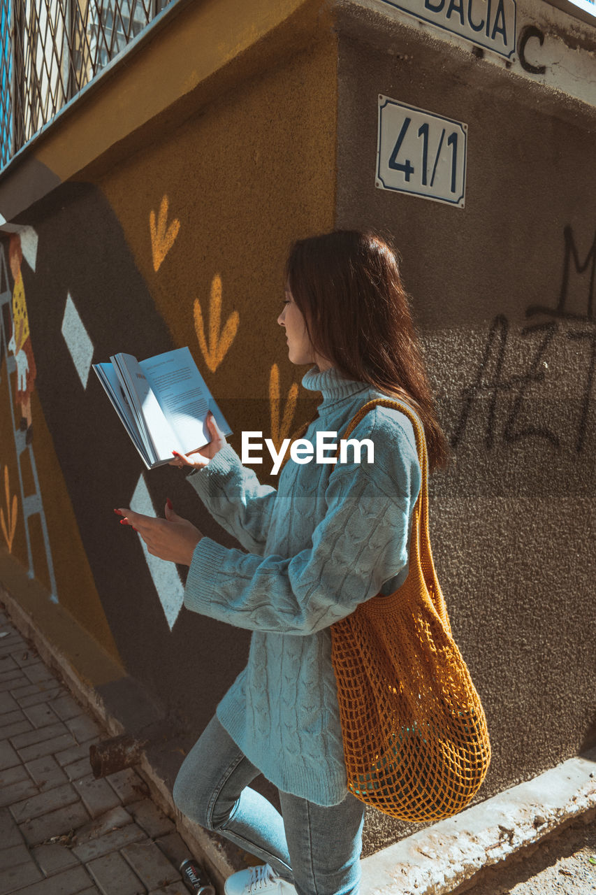 Woman reading book while standing by wall