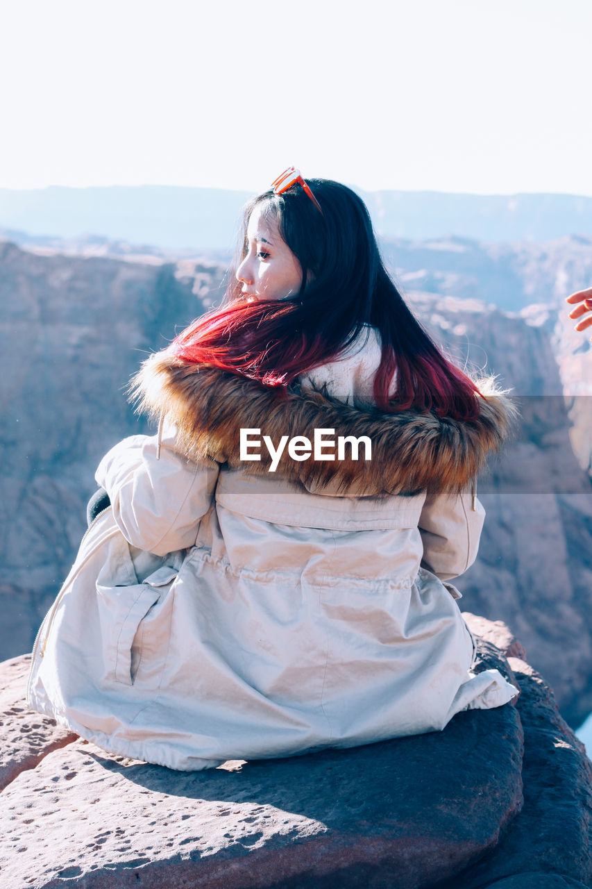 Rear view of woman wearing warm clothing while sitting on cliff