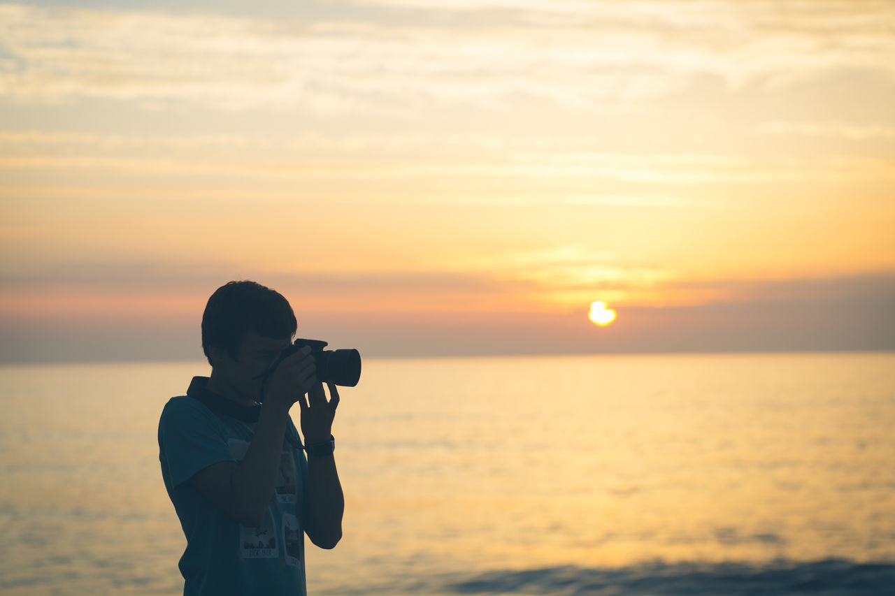 Photographer photographing by sea against sky during sunset