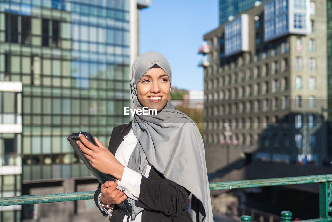 Cheerful muslim female entrepreneur in hijab and with takeaway coffee standing in street