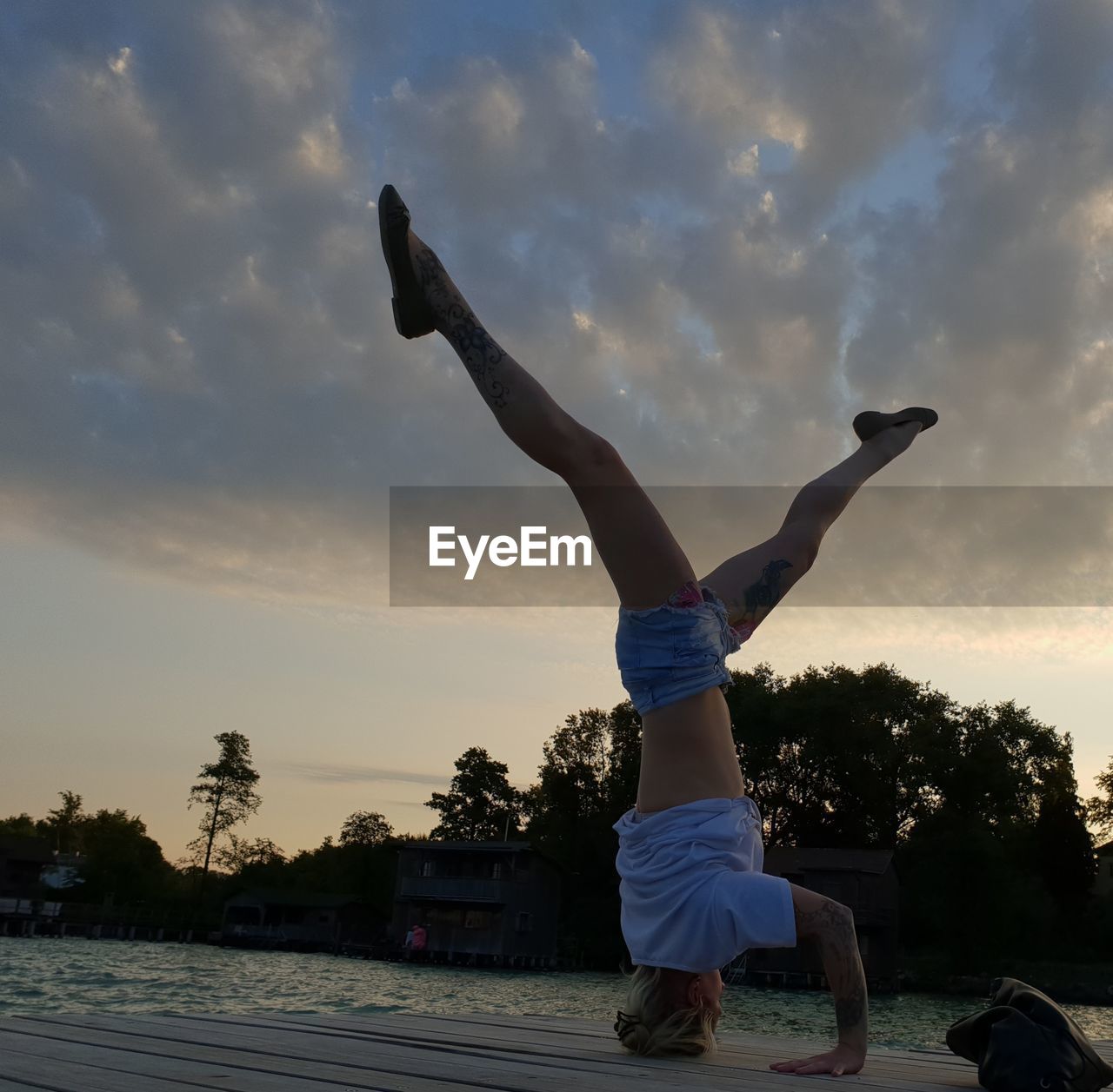 Full length of young woman doing headstand on pier over lake against cloudy sky during sunset