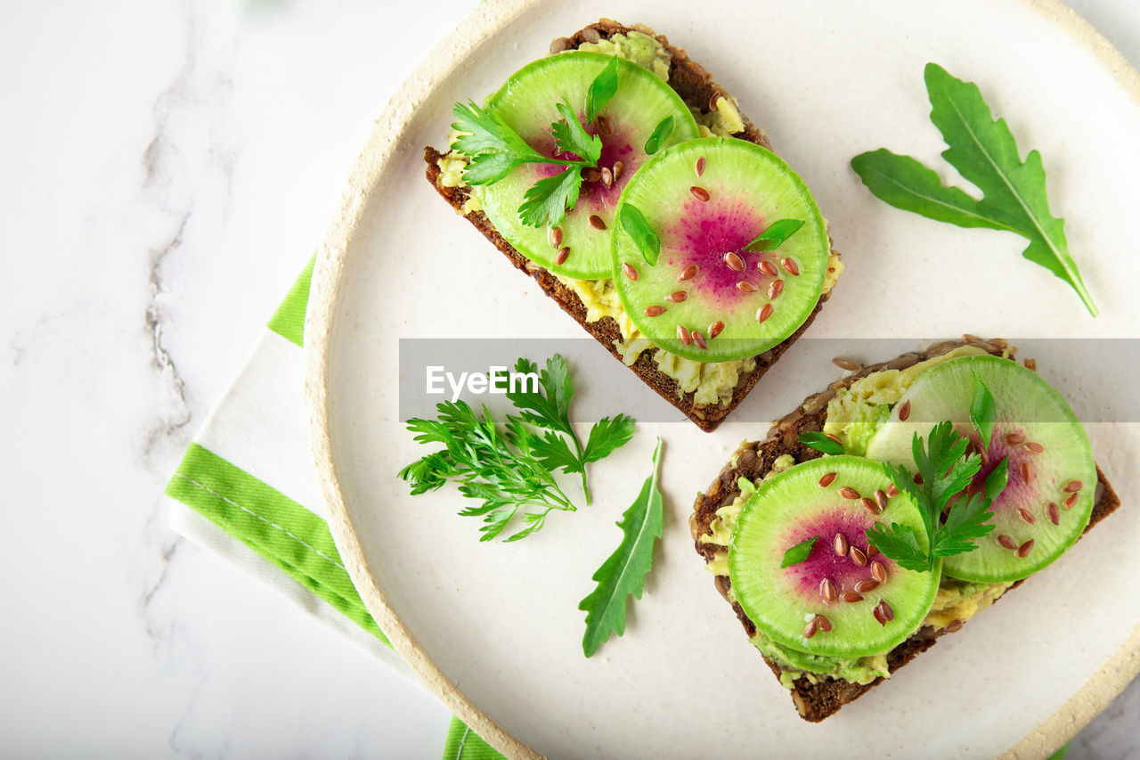 Homemade toasts with organic watemelon radish, avocado and flex seeds on white marble background.