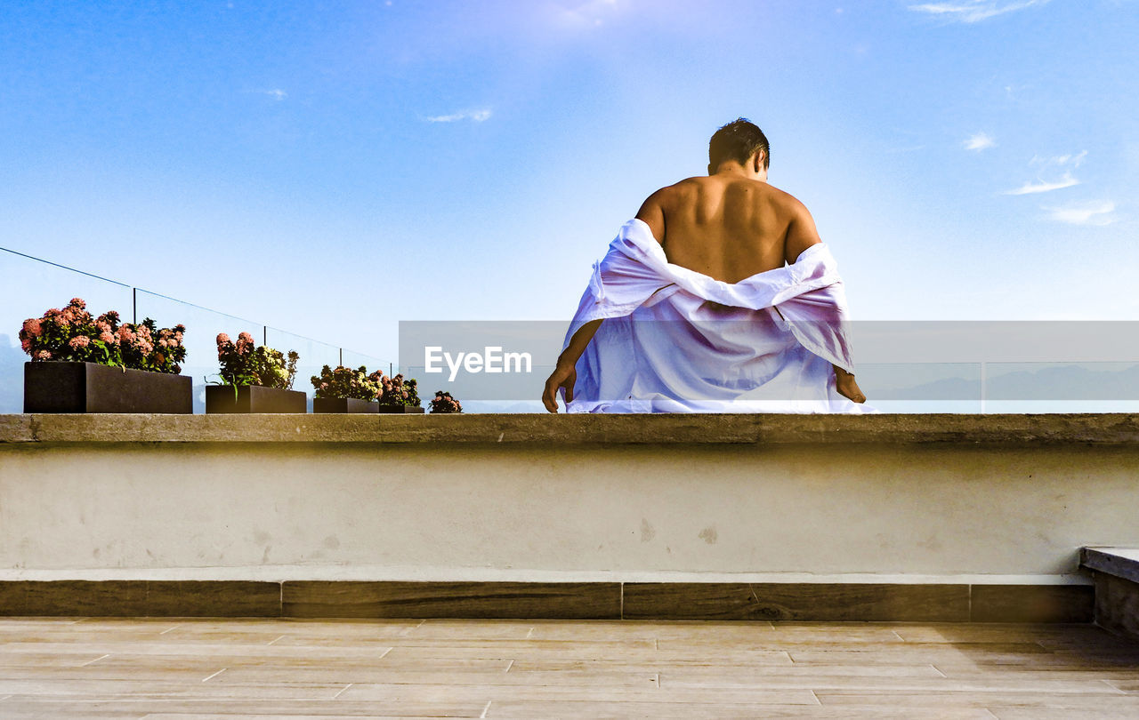 Rear view of shirtless man sitting on retaining wall against sky