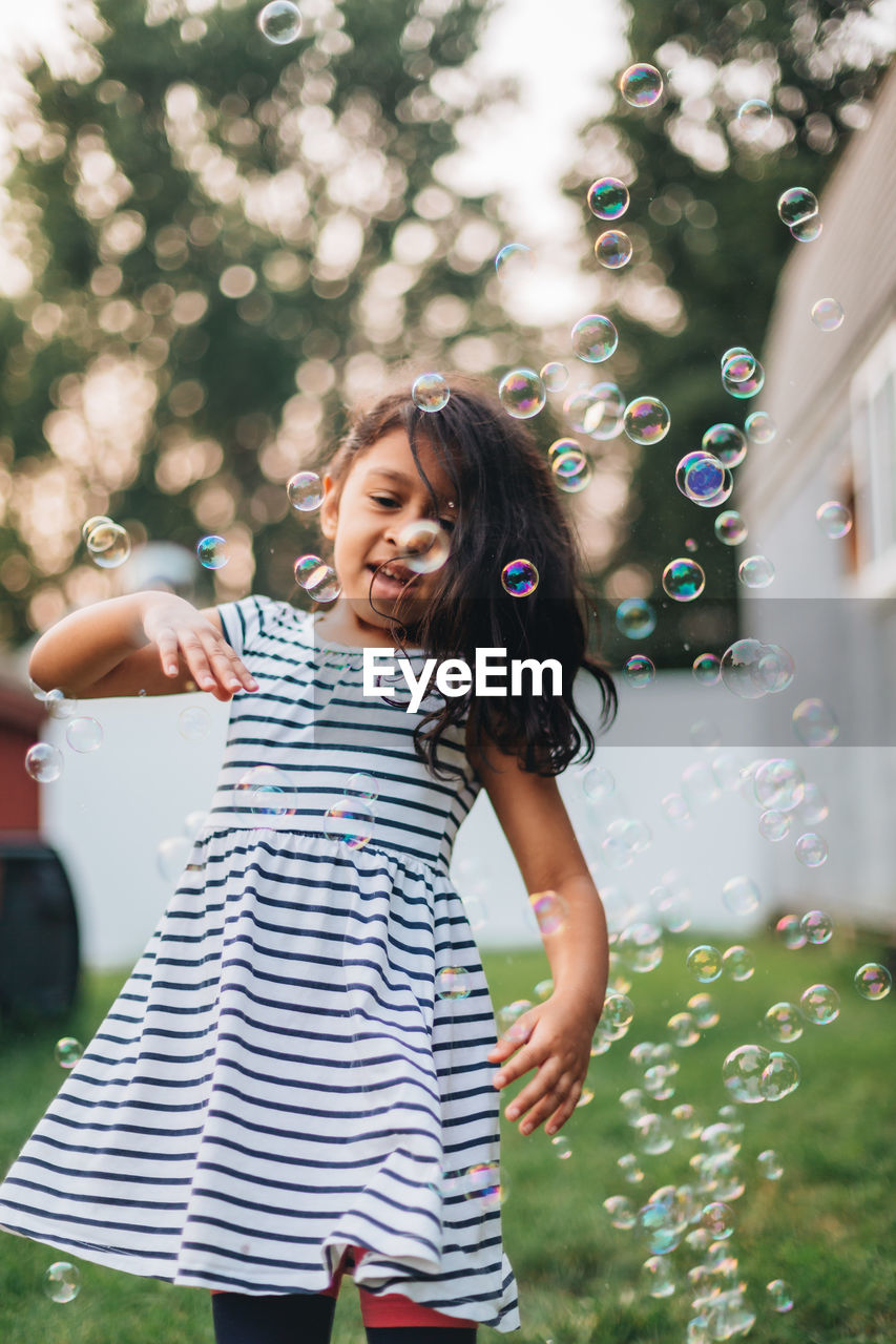 Diverse mixed race pre school girl outdoors during summer having fun in backyard with bubbles 